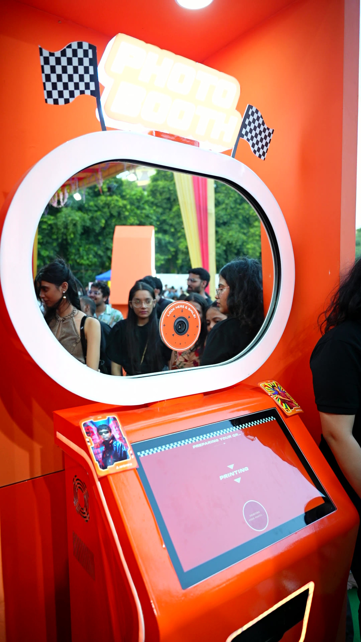 A Pikcha AI photo booth wrapped in a bright orange brand design with a large oval mirror frame and a touchscreen