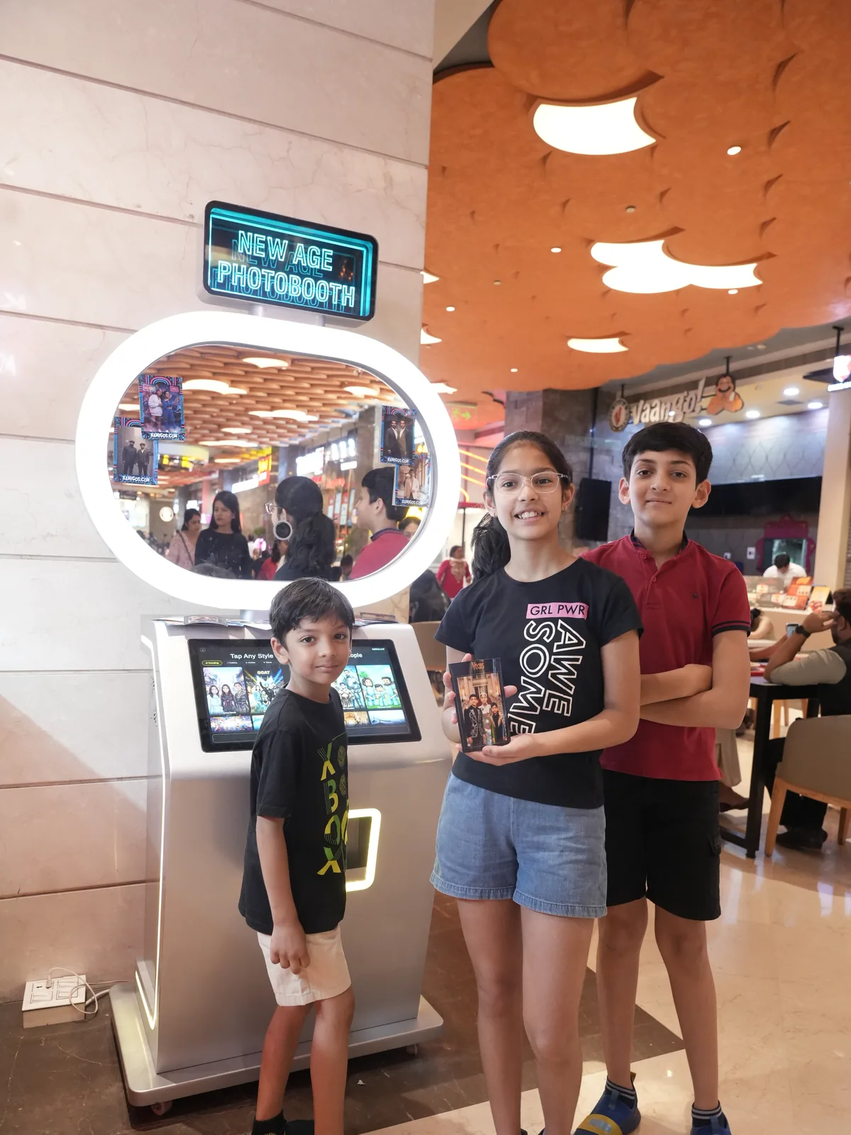 Three siblings holding their instant prints beside the Pikcha photo booth with its glowing ring mirror