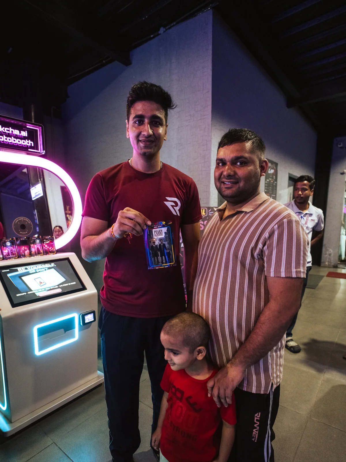 Two men and a child holding a photo booth print beside the Pikcha kiosk in an arcade