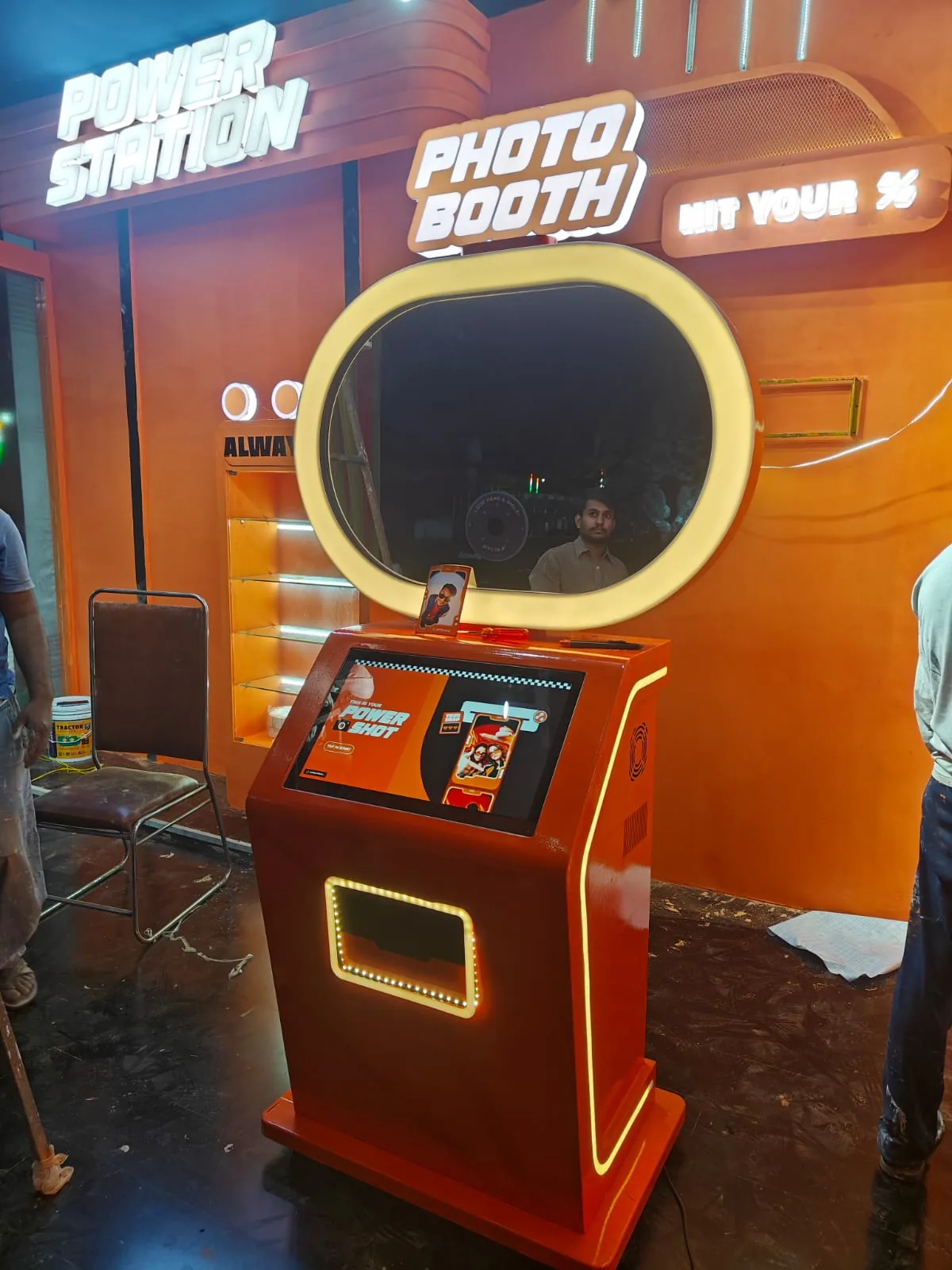 Orange branded Pikcha photo booth installed and lit at night in an event stall, crew still finishing around it